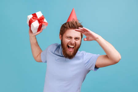 Portrait of excited bearded man in party cone, holding gift box, looks far away with hand over eyes,...
