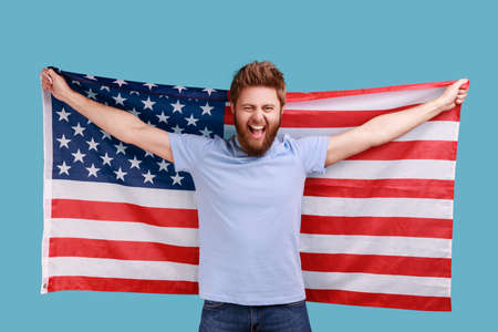Portrait of handsome overjoyed bearded man holding USA flag and looking at camera with rejoicing look, celebrating national holiday. Indoor studio shot isolated on blue background.の写真素材