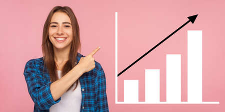 Portrait of happy joyful young woman standing, pointing aside and showing business growth graph. indoor studio shot isolated on pink backgroundの写真素材