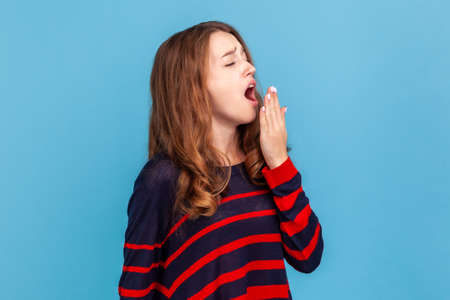 Tired sleepless young adult woman wearing striped casual style sweater, yawning and covering mouth with palm, lack of sleep, fatigue concept. Indoor studio shot isolated on blue background.の写真素材