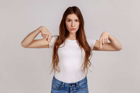 Here and right now. Portrait of woman pointing fingers down, ordering and controlling, demanding result immediately, authority, wearing white T-shirt. Indoor studio shot isolated on gray background.の写真素材