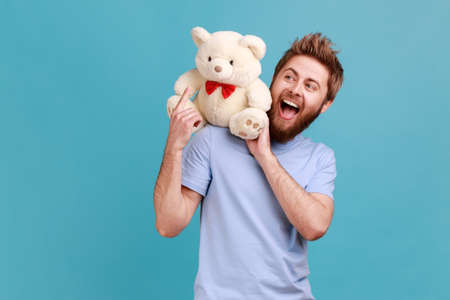 Side view of satisfied delighted handsome bearded man standing, holding white soft teddy bear on his...