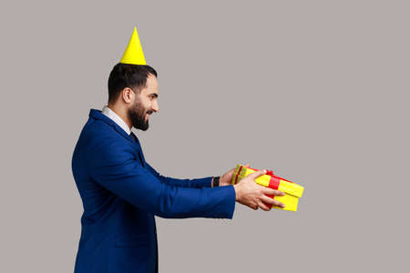 Side view of positive man with festive mood standing in party cone and giving present box, celebrating friends birthday, wearing official style suit. Indoor studio shot isolated on gray background.の写真素材