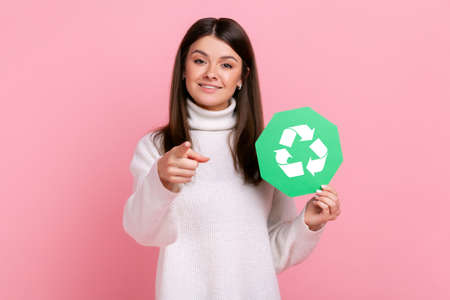 Female holding green recycling sign, pointing to camera, calls on to save our planet from pollution, wearing white casual style sweater. Indoor studio shot isolated on pink background.の写真素材