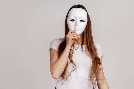 Portrait of anonymous dark haired woman covering her face with white mask, hiding personality,...