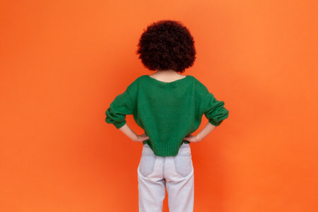 Back view portrait of woman with Afro hairstyle wearing green casual style sweater standing posing backwards, keeping hands on hips. Indoor studio shot isolated on orange background.の写真素材