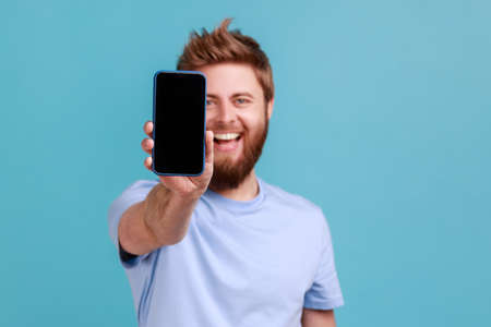 Portrait of bearded man holding out mobile phone and looking at camera with toothy smile and...