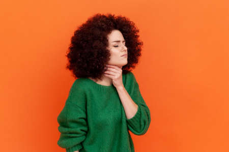 Profile portrait of young adult woman with Afro hairstyle wearing green casual style sweater having sore throat, touching neck, feels bad. Indoor studio shot isolated on orange background.の写真素材