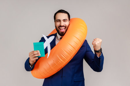 Happy bearded businessman holding orange rubber ring, paper plane and passport, clenching fist, long awaited vacation, wearing official style suit. Indoor studio shot isolated on gray background.の写真素材