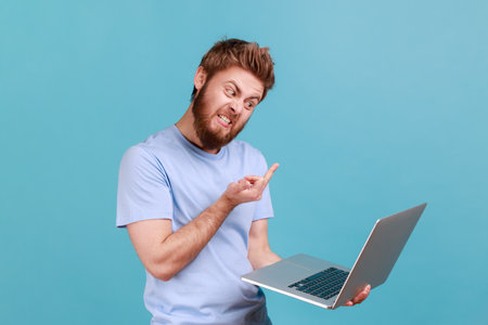 Portrait of bearded man showing middle finger to laptop screen, aggressively communicating on video call, expressing hate to job or coworker. Indoor studio shot isolated on blue background.の写真素材