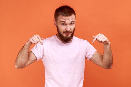 Portrait of man pointing fingers down paying attention at advertising area, showing freespace, looking at camera, wearing pink T-shirt. Indoor studio shot isolated on orange background.の写真素材