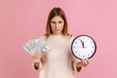 Portrait of upset sad blond woman holding big fan of dollar banknotes and wall clock, time is money, looking at camera, wearing white sweater. Indoor studio shot isolated on pink background.の写真素材