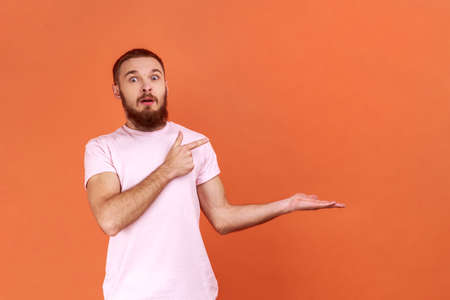 Portrait of surprised bearded man pointing finger right at empty copy space with astonishment, place for advertisement, wearing pink T-shirt. Indoor studio shot isolated on orange background.の写真素材