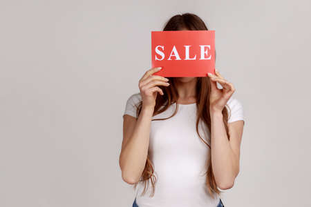 Portrait of anonymous unknown young woman hiding her face behind card with sale inscriptions, shopping, discounts, wearing white T-shirt. Indoor studio shot isolated on gray background.の写真素材