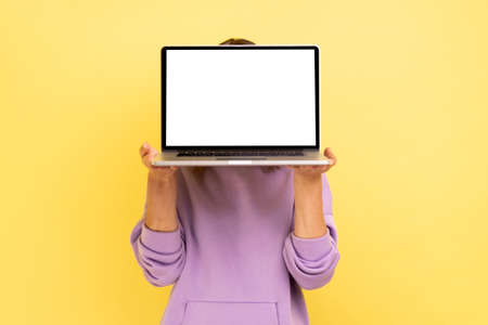 Portrait of unrecognizable woman hiding her face behind laptop with blank display for advertisement...