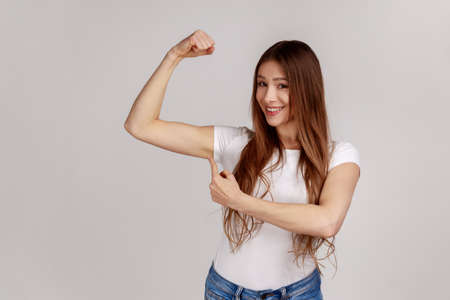 Portrait of confident independent woman raising hand pointing biceps, feeling power and energy to achieve goal, wearing white T-shirt. Indoor studio shot isolated on gray background.の写真素材