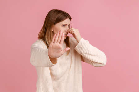 Portrait of woman pinching her nose, holding breath to avoid bad smell and showing stop gesture,...