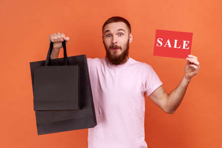 Portrait of man posing with shopping bags and card with sale inscription in hands, looking at camera with surprised expression, wearing pink T-shirt. Indoor studio shot isolated on orange background.の写真素材