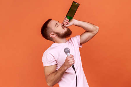 Portrait of drunk bearded man spending time in karaoke, holding microphone in hand, drinking alcoholic beverage, wearing pink T-shirt. Indoor studio shot isolated on orange background.の写真素材