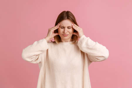 Portrait of unhealthy sick blond woman massaging temples with hands feeling terrible headache, risk of migraine, wearing white sweater. Indoor studio shot isolated on pink background.の写真素材