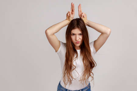 Portrait of aggressive bully woman with dark hair showing bull horns gesture over head, frowning as...