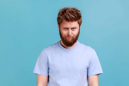 Portrait of bearded upset young man standing and looking at camera with dissatisfied sadness face,...