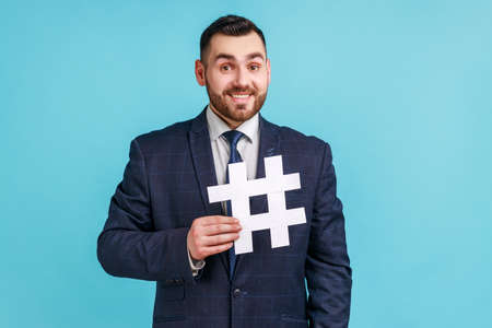 Smiling businessman wearing dark official style suit holding hashtag symbol and looking at camera...