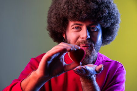 Portrait of hipster man with Afro hairstyle holding red toy heart, showing his fondness and devotion, romance, wearing red sweatshirt. Indoor studio shot isolated on colorful neon light background.の写真素材