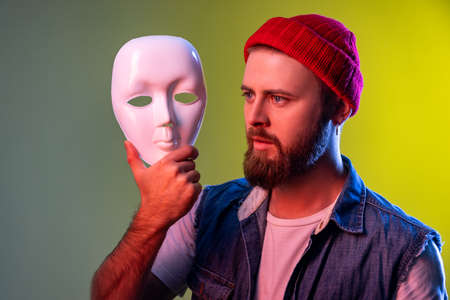 Confident serious hipster man holding white mask in hands, looking away with attentive look, hiding personality, wearing beanie hat. Indoor studio shot isolated on colorful neon light background.の写真素材