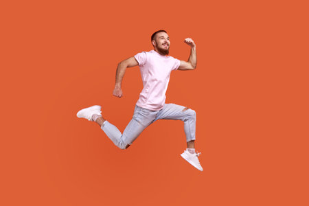 Full length portrait of positive inspired bearded man jumping in air or running quickly fast, expressing happiness, wearing pink T-shirt. Indoor studio shot isolated on orange background.の写真素材