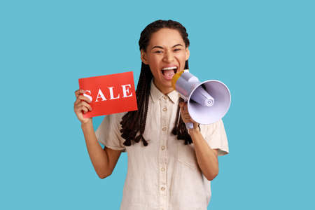 Portrait of emotional woman with black dreadlocks holding card with sale inscription and screaming in megaphone about discounts, wearing white shirt. Indoor studio shot isolated on blue background.の写真素材