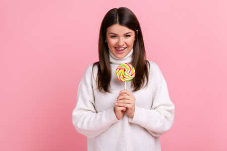 Portrait of hungry brunette female holding big tasty candy, looking at camera, showing tongue out, wearing white casual style sweater. Indoor studio shot isolated on pink background.の写真素材