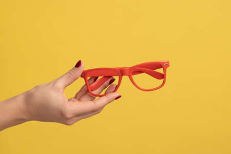 Profile side view closeup of woman hand holding and showing red eyeglasses frame. Indoor studio shot isolated on yellow background.の写真素材