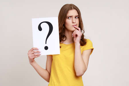 Young pensive woman in casual style attire holding paper with question mark over, thoughtful, face thinking about question, very confused idea. Indoor studio shot isolated on gray background.の写真素材