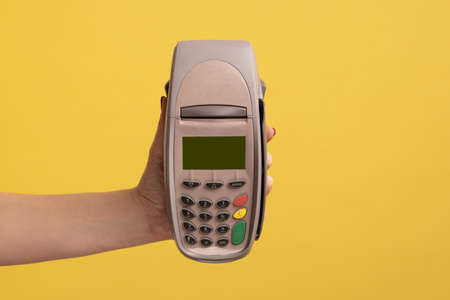 Profile side view closeup of woman hand holding and showing POS or terminal payment or pay pass device. Indoor studio shot isolated on yellow background.の写真素材