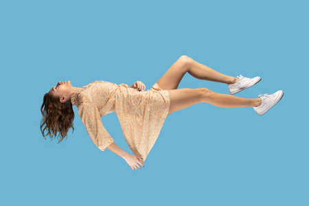 Floating in air. Relaxed girl in yellow dress levitating keeping eyes closed, sleeping while flying mid-air, having comfortable peaceful dream. full length studio shot isolated on blue background, indoorの写真素材