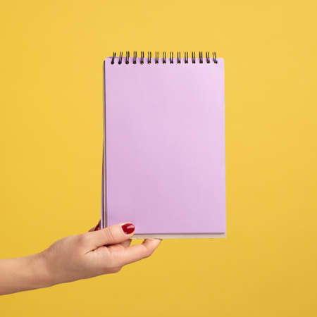 Profile side view closeup of woman hand holding purple notepad in hand and showing empty paper. Indoor studio shot isolated on yellow background.の写真素材