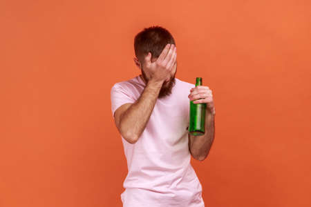 Portrait of upset sad bearded man standing with beer bottle, being drunk, standing covering his face...