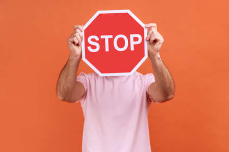 Portrait of man covering face with Stop symbol, anonymous person holding red traffic sign, warning to go, prohibition concept, wearing pink T-shirt. Indoor studio shot isolated on orange background.の写真素材