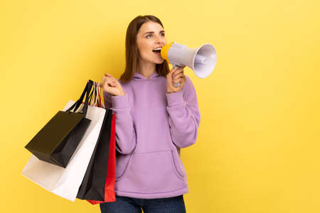 Young beautiful woman with brown wavy hair holding shopping bags and megaphone, announcing sale and discount, wearing purple hoodie. Indoor studio shot isolated on yellow background.の写真素材