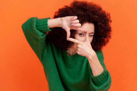 Young adult woman with Afro hairstyle wearing green casual style sweater standing crop composition focus hand gesture and looking at camera. Indoor studio shot isolated on orange background.の写真素材
