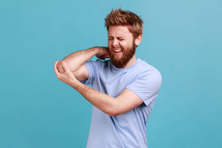 Portrait of bearded man massaging aching elbow, frowning from acute pain, strain and strain problem, trapped nerves, rheumatoid arthritis. Indoor studio shot isolated on blue background.の写真素材