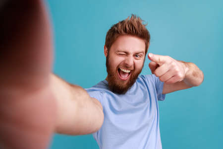 Portrait of satisfied delighted bearded man blogger pointing at you, looking at camera with toothy smile, POV, point of view of photo. Indoor studio shot isolated on blue background.の写真素材