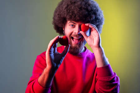 Hipster man with Afro hairstyle having fun with red toy hearts, showing his fondness and devotion, romance, wearing red sweatshirt. Indoor studio shot isolated on colorful neon light background.の写真素材