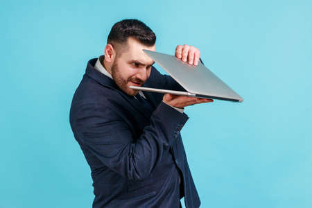 Portrait of curious bearded businessman wearing official style suit carefully peeking into half-opened laptop and spying on forbidden content. Indoor studio shot isolated on blue background.の写真素材