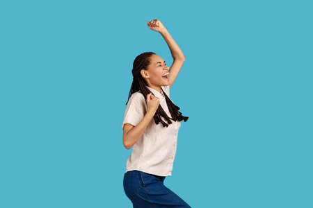 Portrait of excited woman with black dreadlocks clenches fists, celebrates success, feels happy after winning or triumph, wearing white shirt. Indoor studio shot isolated on blue background.の写真素材