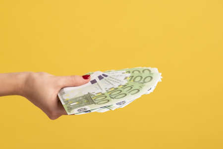 Closeup side view female hand holding and giving fan of euro banknotes, big salary, profit, lot of money and wealthy life. Indoor studio shot isolated on yellow background.の写真素材