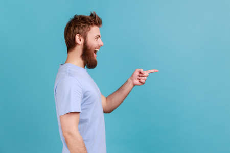Side view of positive satisfied bearded man pointing aside, showing blank copy space for idea presentation, commercial text, smiling happily. Indoor studio shot isolated on blue background.の写真素材