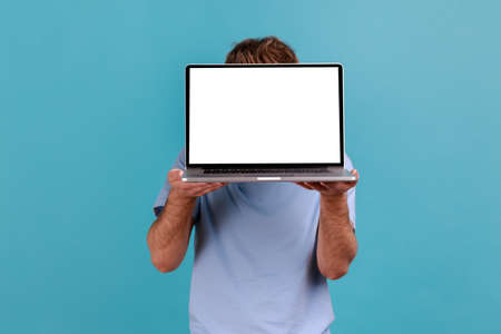 Portrait of unknown man hiding face behind laptop with white empty display and presenting new...