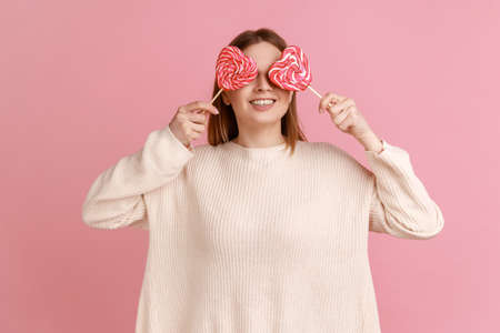 Portrait of satisfied smiling blond woman covering her eyes with tasty heart shape lollypops, having...